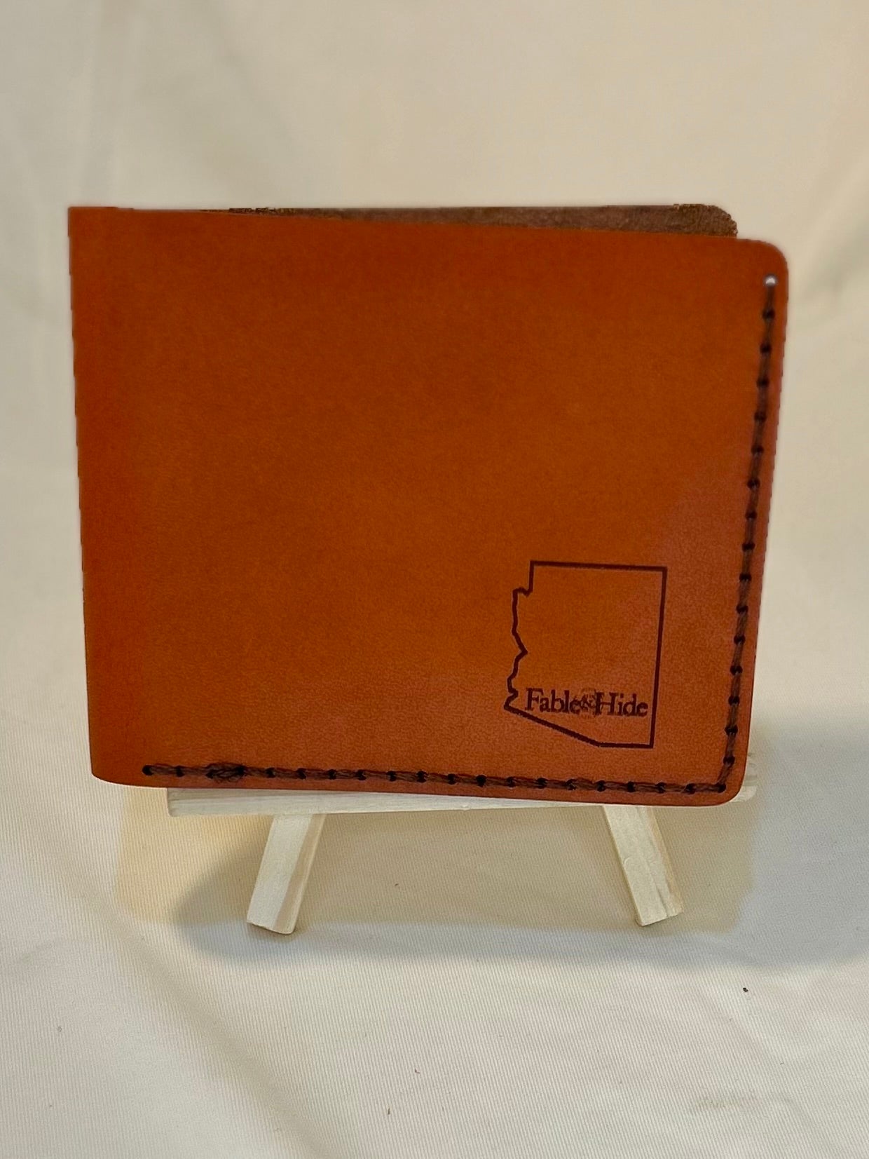 Outside of an orange wallet. There is an engraving of an outline of the state of Arizona with a Fable&Hide makers mark inside and brown stitching on the outside 