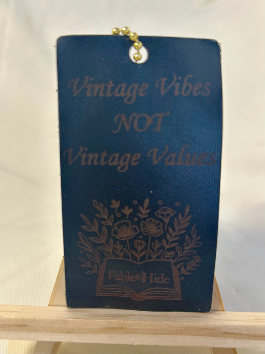 Black rectangle leather charm with gold ball chain. The charm says “Vintage Vibes NOT Vintage Values” and has an open book with flowers image with the Fable&Hide makers mark on the book pages 